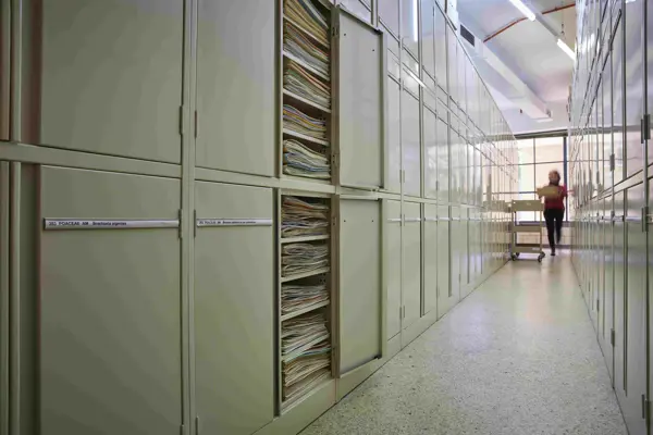 Two rows of metal cream-coloured metal cupboards, with thin paper labels on each door. The doors of two cabinets are open, with paper folders showing inside. At the far end of the aisle, a blurry person carrying a pile of folders approaches a trolley.