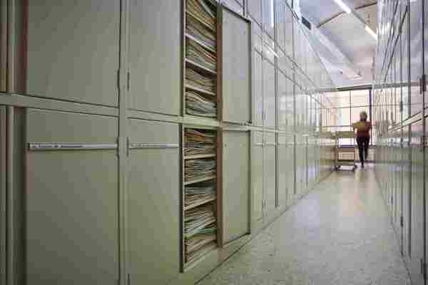 Two rows of metal cream-coloured metal cupboards, with thin paper labels on each door. The doors of two cabinets are open, with paper folders showing inside. At the far end of the aisle, a blurry person carrying a pile of folders approaches a trolley.