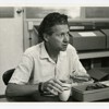 A black and white, candid photo of Rex Filson. He is eating lunch at a table by a shelf of boxed archival materials.