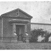 A black and white photo of a grey, stone building on a sparse lawn. A square archway leads to a door at the front. Two people pose on the step under the archway. The building continues to the right with a few trees obscuring several windows.