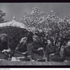 A black and white photo of five women sitting on a lawn beneath a blossoming tree, eating lunch and looking at one another. They are all wearing a uniform of a skirt and jacket. A roof with a decorative spike at its apex is visible in the background.