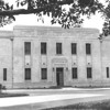 A black and white photo of a building taken from a vantage point across the street under a tree with low branches. The building is very square, with decorative motifs along the top and several columns of rectangular windows over two storeys. The door to the building resides within a square archway with a lantern either side.